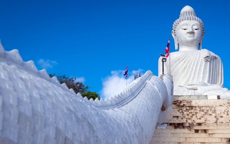 Majestic Big Buddha statue in Phuket, Thailand, with white staircase and Thai flags under a clear blue sky
