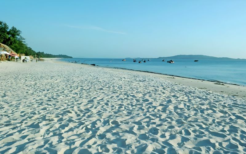 A wide stretch of white sandy beach with gentle waves, small fishing boats floating offshore, and a clear blue sky in a quiet coastal setting.