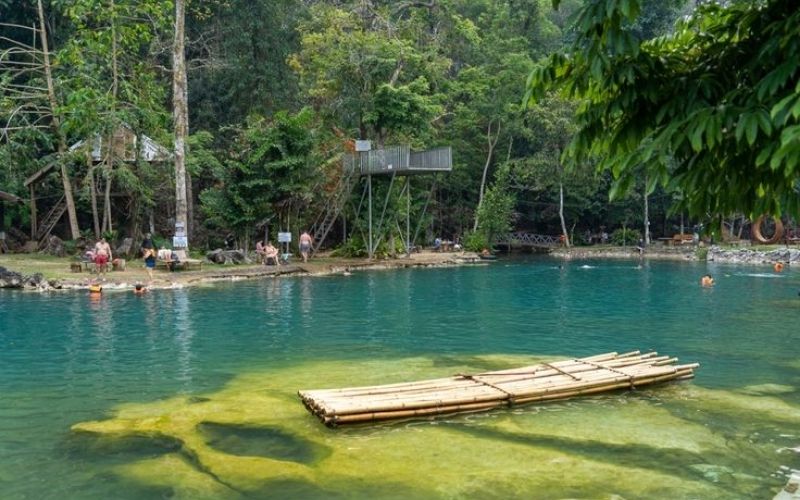 Scenic lagoon with a wooden raft surrounded by lush green trees in Vang Vieng, Laos.