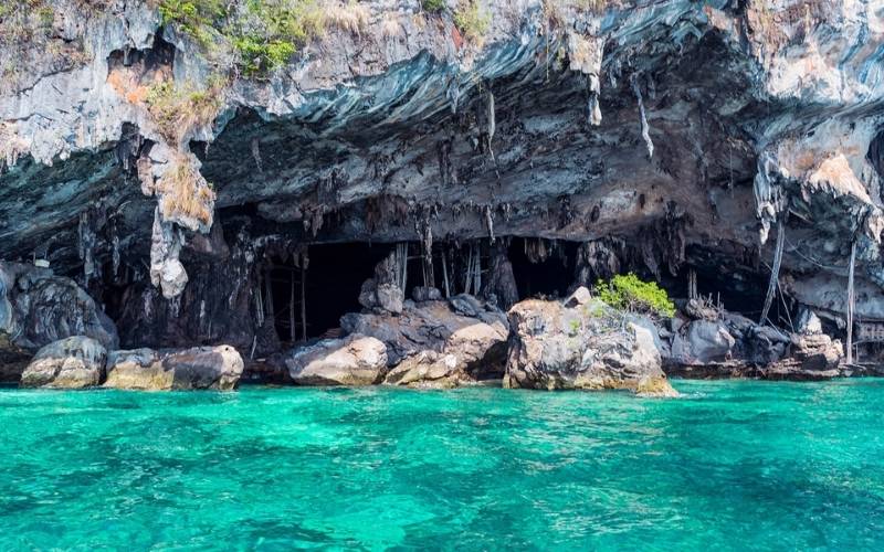 A striking sea cave with intricate rock formations and stalactites hanging from its entrance, situated above vibrant turquoise water. Large boulders are visible in front of the cave, with lush greenery growing on the cliff tops.