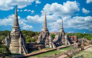 Ancient temple ruins with three large chedis at Wat Phra Si Sanphet in Ayutthaya, Thailand, under a bright blue sky with scattered clouds