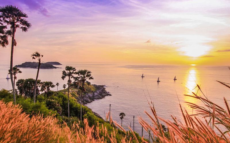 Scenic sunset view at Promthep Cape in Phuket, Thailand, with golden sky, ocean horizon, and silhouetted palm trees