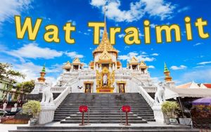 A wide-angle, vibrant view of the white and gold Phra Maha Mondop building at Wat Traimit in Bangkok, featuring broad stone stairs leading up to the temple under a bright blue sky with scattered clouds. The words "Wat Traimit" are overlaid in large yellow letters across the top.