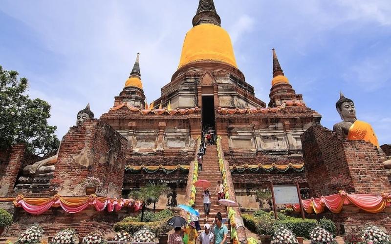 A low-angle view of the massive central chedi at Wat Yai Chai Mongkhon in Ayutthaya, decorated with yellow robes and festive pink bunting, as visitors climb the steep staircase to the entrance.