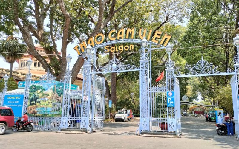 A wide view of the historic main entrance gate of the Saigon Zoo and Botanical Gardens (Thảo Cầm Viên) on Nguyen Binh Khiem Street, featuring three arched walkways, a red-tiled roof, and vibrant red and yellow signage against a backdrop of lush green trees.