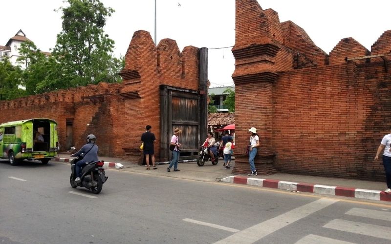 A close-up view of the historic red brick walls and wooden gate of Tha Phae Gate in Chiang Mai, with traffic (a motorbike and a green tuk-tuk) moving past the foreground.