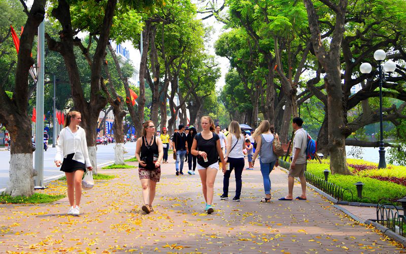 Tourists walking along the tree-lined pedestrian path of the Hoan Kiem Lake walking street in Hanoi, with fallen yellow leaves scattered on the ground and red flags visible in the background.