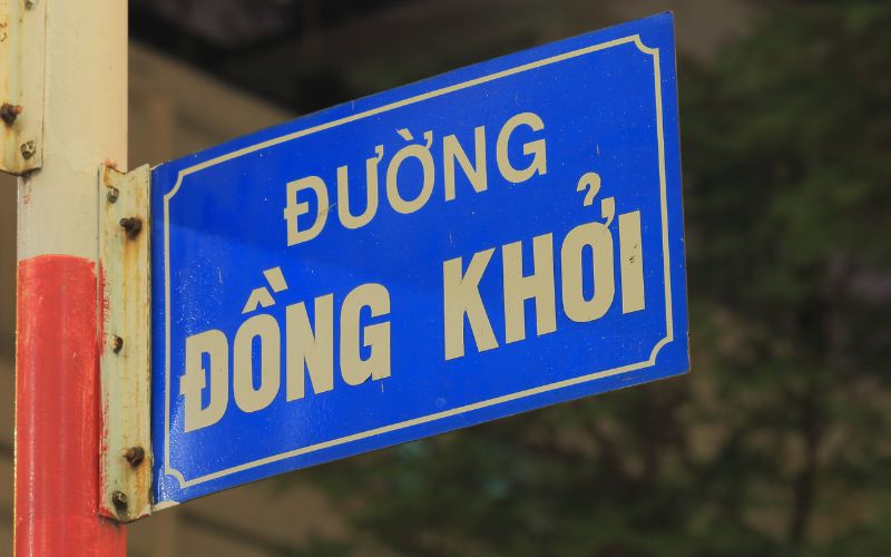 A close-up of a blue street sign with white lettering that reads "ĐƯỜNG ĐỒNG KHỞI" against a soft-focus background of a white building.