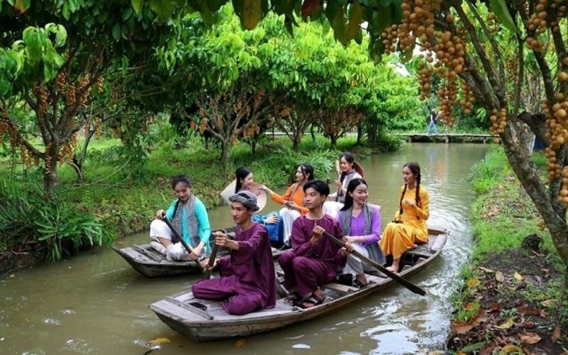 A group of young Vietnamese people in traditional silk outfits paddle a wooden boat through a garden canal. Longan fruit clusters hang from the trees directly above the water as the group smiles and navigates through the orchard during the River Culture Festival.