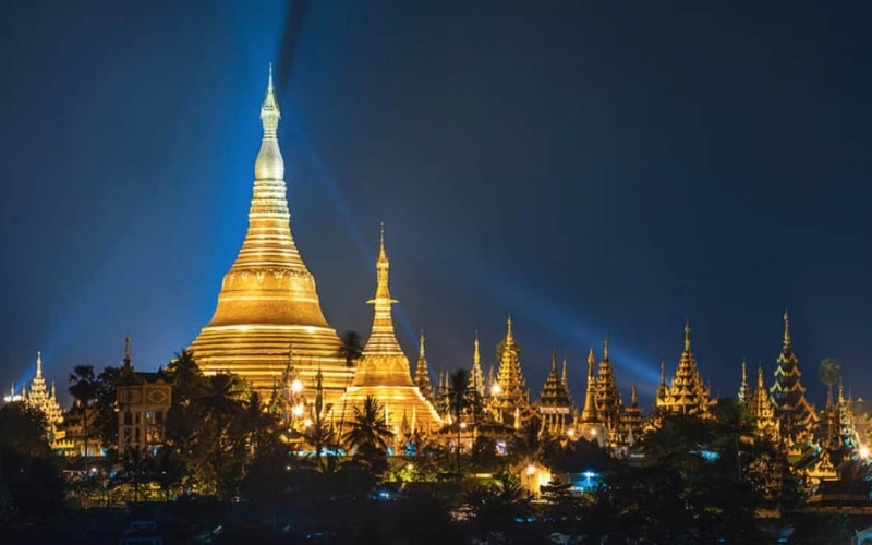 The golden Shwedagon Pagoda in Yangon, Myanmar, illuminated brightly against the night sky with lights reflecting off its golden spires.