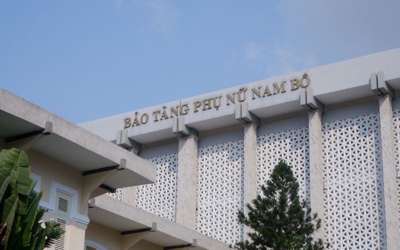 The exterior of the Southern Women's Museum in Ho Chi Minh City, featuring a large, multi-story white building with modern architectural lines. A prominent red banner with Vietnamese text hangs above the entrance, and the museum is surrounded by lush green trees and a paved courtyard.