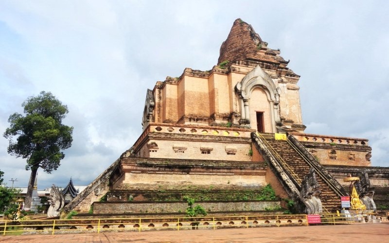 An outdoor wide shot of the ancient, partially ruined main chedi (stupa) of Wat Chedi Luang in Chiang Mai, Thailand. The large, weathered brick structure shows signs of age and earthquake damage, with steep stone staircases leading up its sides and Nāga sculptures guarding the lower level. A single tall, leafy green tree stands to the left of the chedi against a bright but cloudy sky.