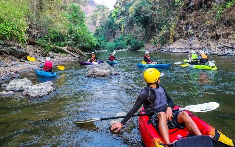 A group of people wearing helmets and life jackets paddling colorful kayaks down the rocky Mae Taeng River near Chiang Mai, surrounded by dense jungle.
