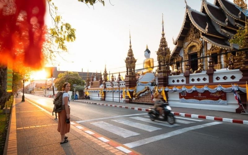 A female tourist with a backpack stands on a sidewalk, looking towards an ornate Thai temple featuring a large seated Buddha statue, illuminated by the golden light of sunrise or sunset in Chiang Mai.