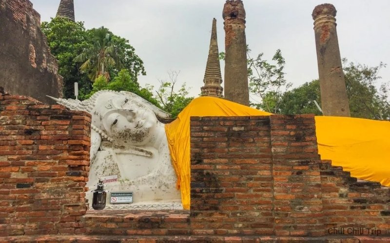A large white reclining Buddha statue draped in bright yellow robes, situated behind ancient brick walls and crumbling columns at Wat Yai Chai Mongkhon.