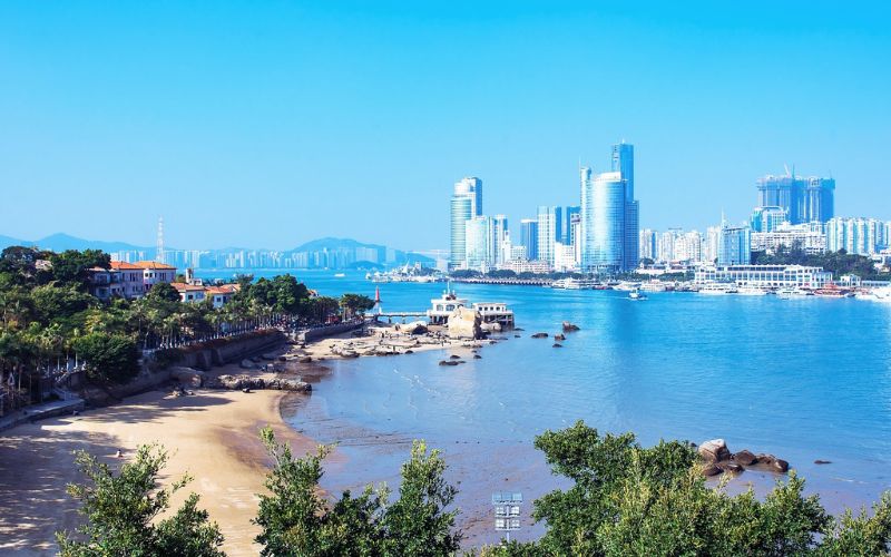 A bright and clear daytime view of Xiamen cityscape featuring modern high-rise buildings along the coastline, with a sandy beach, lush greenery in the foreground, and calm blue waters under a clear blue sky.