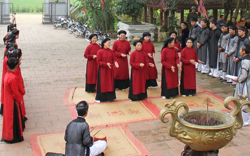 Group of performers in traditional red áo dài singing Xoan folk songs in a courtyard, with musicians seated and villagers watching in the background