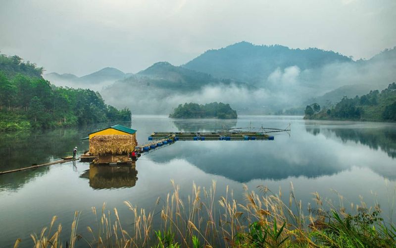 Tranquil lake scene at Xuan Son National Park in Phu Tho, Vietnam, with a thatched hut on the water, misty mountains, and lush greenery in the background