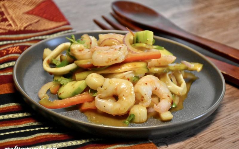 Plate of Thai yum talay seafood salad served on a dark round dish, featuring shrimp, squid rings, sliced onion, tomato, celery, herbs, and a spicy tangy dressing, set on a wooden table with Thai-patterned fabric and wooden utensils in the background.